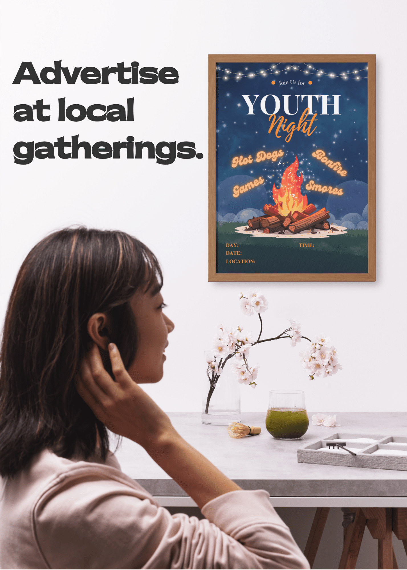 A young adult woman notices a framed poster of 'Youth Bonfire Night' with a campfire design on the wall.