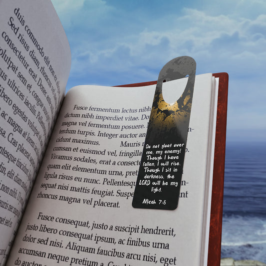 The Bible verse page marker "Don't Gloat over me", inspired by Micah 7:8, is clipped securely to a book page and held in front of a soft beach background. A Christian bookmark made for Bible study, devotionals, or gifting—mark the moment you didn’t quit trusting Him.